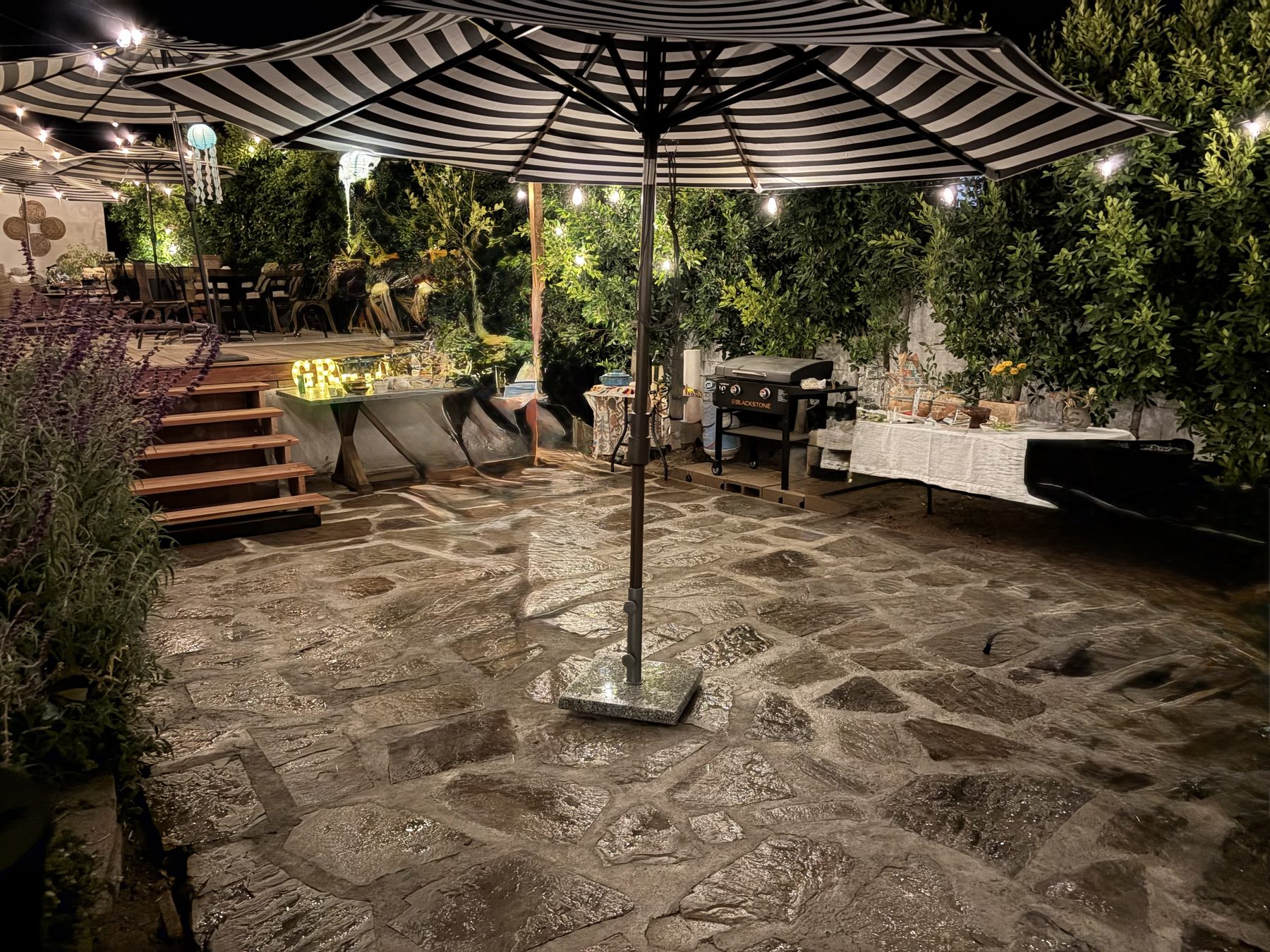 Flagstone courtyard at night under a black-and-white striped umbrella, string lights and greenery all around